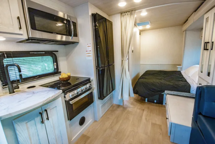 Inside this RV rental: a compact kitchen with microwave, stove, sink, and a large fridge adjacent to a cozy bedroom.. Nexus Other 2026