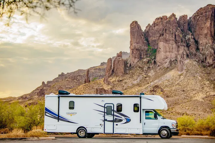 A white RV rental with blue graphics is parked on pavement, facing towering desert mountains under a warm, cloudy sky.. Nexus Other 2026