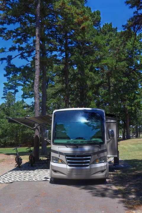 A modern RV rental is parked at a scenic campsite, featuring an extended awning, bikes, and a cozy outdoor rug amidst tall pine trees.. Thor Axis 2015