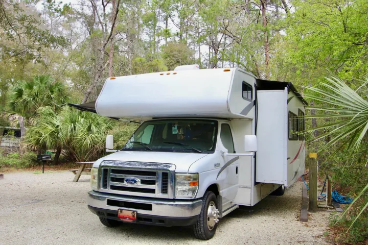 White Class C RV rental parked on a gravel campsite, with picnic table & grill nearby under trees.. Forest River Coachmen Freelander 2011