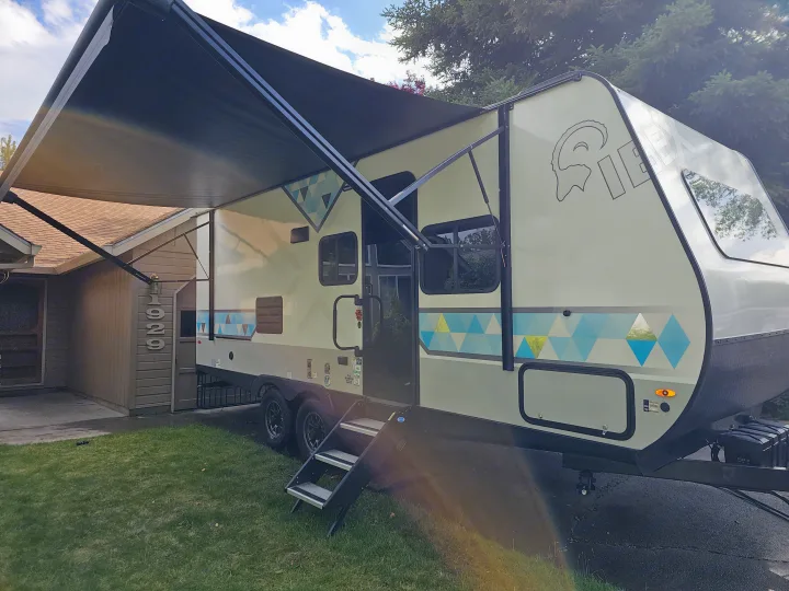 A cream-colored IBEX travel trailer with blue and yellow geometric designs, its dark awning extended, is parked for an RV rental.. Forest River Ibex 2024