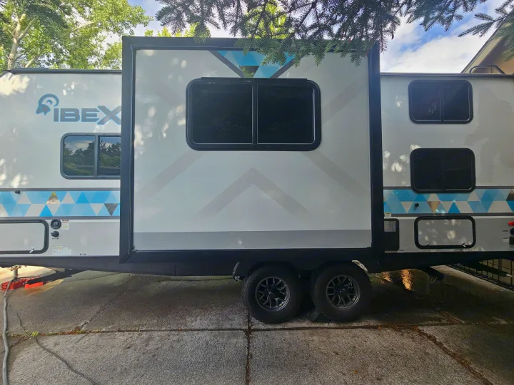 This IBEX travel trailer, a great option for an rv rental, is parked on concrete under trees, showcasing its slide-out and dual axles.. Forest River Ibex 2024