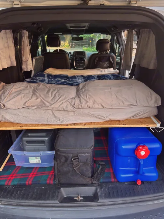 Inside an RV rental: a cozy van camping setup with a portable bed platform, storage bin, cooler, and water jug. Ready for adventure!. Dodge Dodge Grand Caravan GT 2017 2017