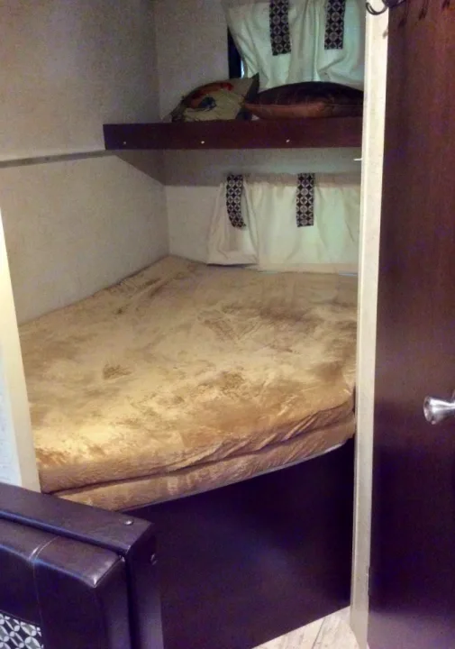 RV rental sleeping area: A lower bunk bed with a soft blanket, and an upper shelf with pillows and patterned curtains.. kz 26' SPORTSMAN 2018