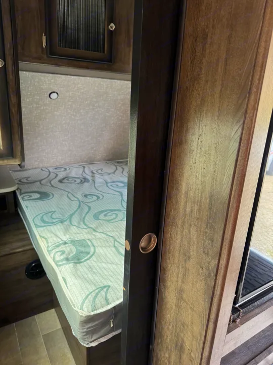 Interior of an RV bedroom showing a bed with a patterned mattress, overhead cabinetry, and a partially open sliding door. Ideal for an RV rental.. Heartland RVs Mallard 2019