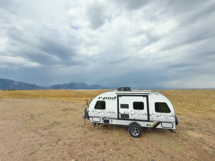 A white r-pod RV rental with a honeycomb pattern is parked in a vast, dry field under a dark, cloudy sky, with mountains in the distance.. R-Pod R-Pod Trailer 2021