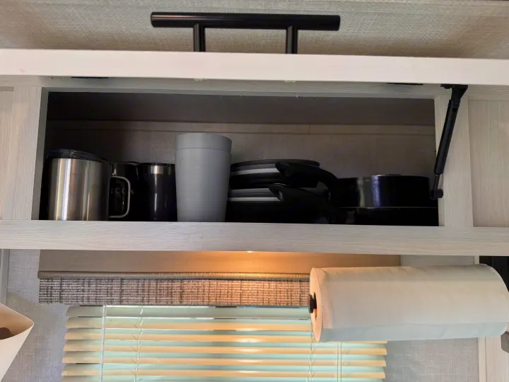 RV rental kitchen shows open overhead cabinet with stacked mugs, cups, plates, and nested pans. Paper towel roll below. Efficient storage.. R-Pod R-Pod Trailer 2021
