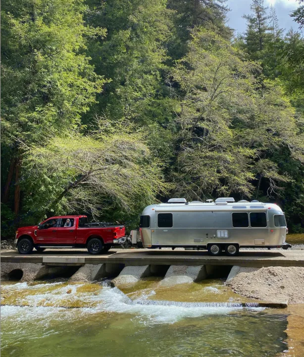 An RV rental adventure: Red truck towing a silver Airstream travel trailer over a rushing river, surrounded by green trees.. Airstream Globetrotter 2019