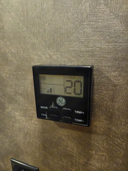 Black GE digital thermostat in an RV rental, set to 20°C (cool mode), fan on. Against a textured grey wall.. Forester LE 2023