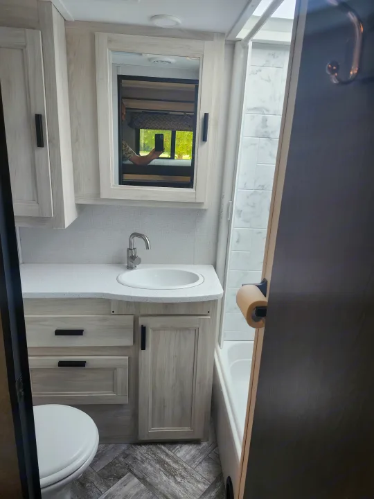 Bright RV rental bathroom featuring a light wood vanity, sink, toilet, and a tiled shower area, with modern black hardware.. Forester LE 2023