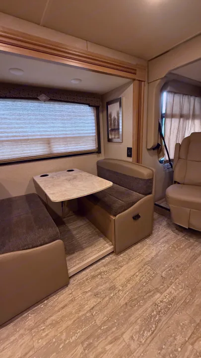 RV rental interior featuring a dinette with a light-colored table, comfortable upholstered seating, and a large window with blinds.. Thor Hurricane 2019
