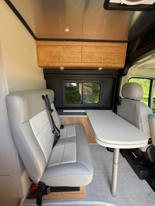 Modern RV rental interior with light gray seating, dining table, and wood cabinetry, offering a scenic window view.. Airstream Airstream Class B 2023