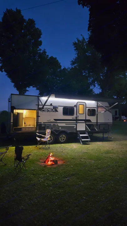 Cozy RV rental at dusk, with an Apex Ultralite camper lit up, featuring an open kitchen, chairs, and a warm campfire.. Coachmen Apex 2023