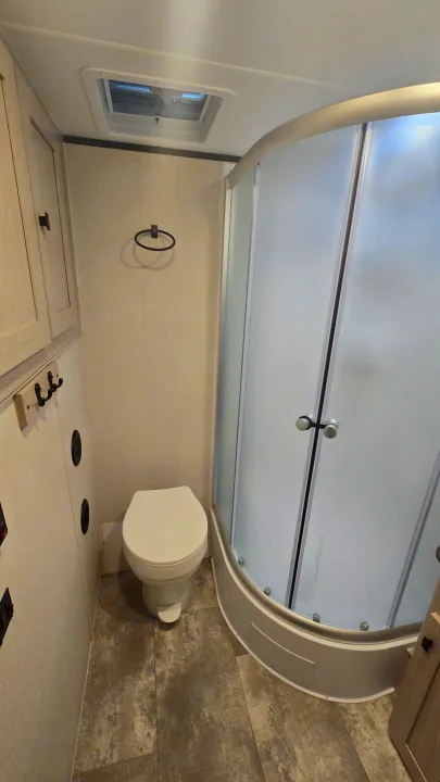 Inside an RV rental: a modern, compact bathroom featuring a toilet, corner shower with frosted glass, and ceiling vent.. Coachmen Apex 2023