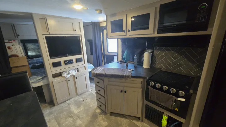 Modern rv rental interior: compact kitchen (stove, microwave, sink), TV, and a glimpse of the bedroom.. Coachmen Apex 2023