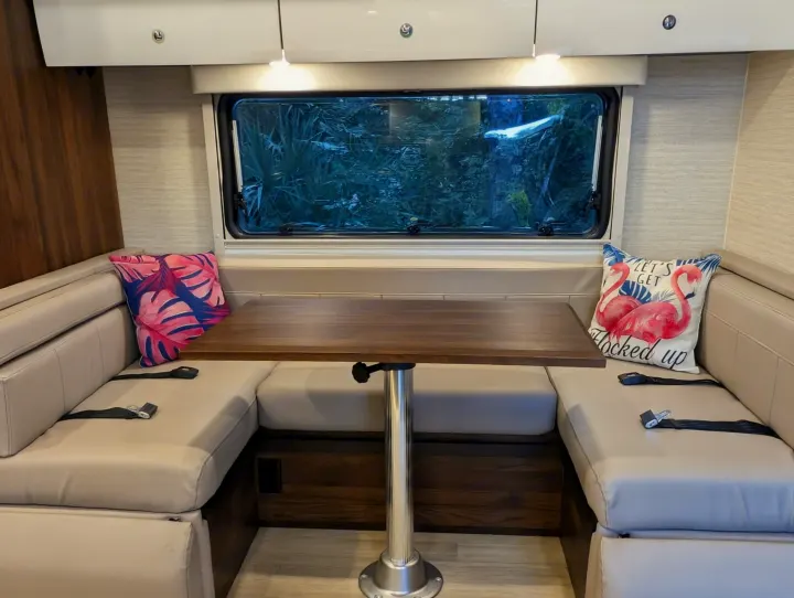 An RV rental's U-shaped dinette with light cushions, a wooden table, and a window framing dense greenery, plus fun pillows.. Winnebago View 2025