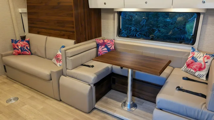 Modern RV rental interior with comfortable U-shaped seating, a dining table, and a large window showing green foliage.. Winnebago View 2025