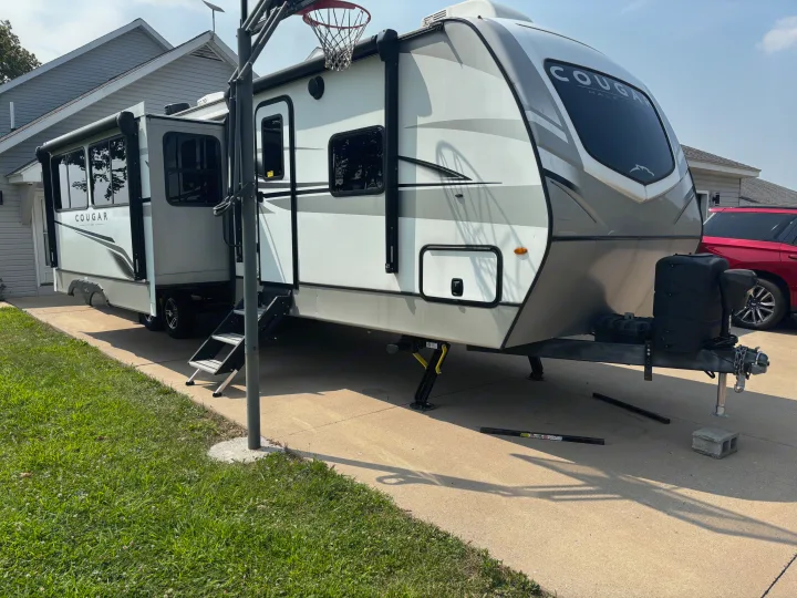 A white Cougar RV rental is set up on a residential driveway, with its side entry steps extended.. Keystone RV Cougar 2023