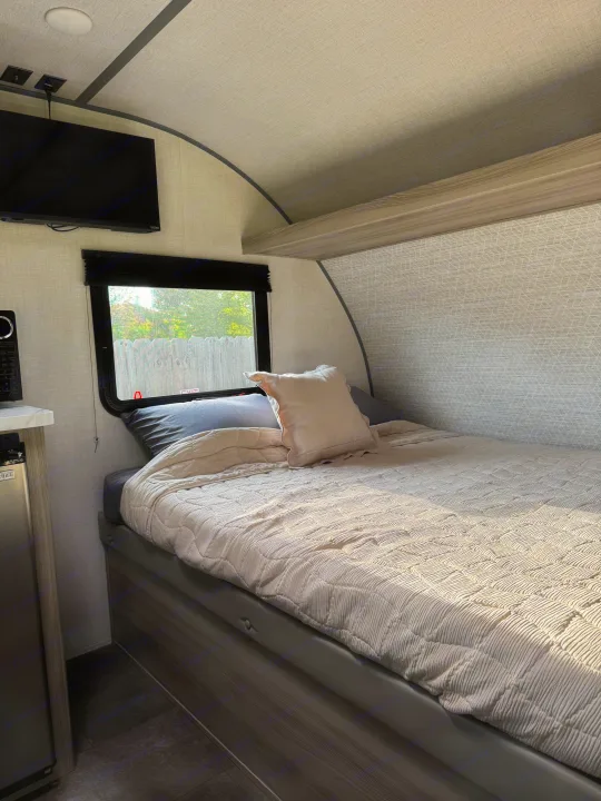Inside a modern rv rental: a cozy bedroom with a quilted bed, mounted TV, window view, and mini-kitchen.. Forest River Wildwood FSX Platinum 2025