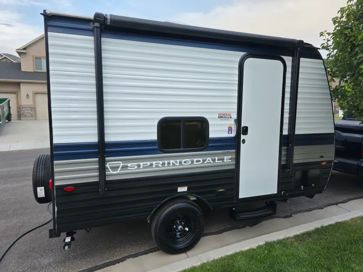 A new, compact travel trailer with blue/grey stripes and an awning, ideal for an rv rental, is parked curbside in a residential area.. Keystone Springdale 2026