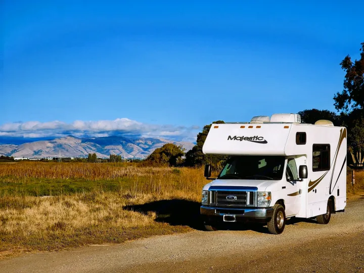 White Majestic Class C motorhome parked on a gravel lot with tall grassy marshland and rolling hills in the background under a clear blue sky — bright, open setting ideal for scenic road trips.. Thor Majestic 2017