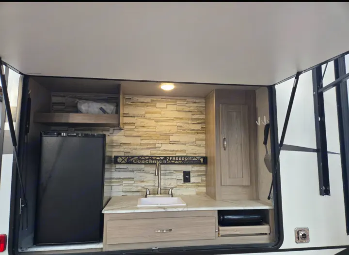 Explore this RV rental's exterior kitchen, equipped with a mini-fridge, sink, and pull-out cooktop on a countertop with a stone backsplash.. Coachmen Forest River Freedom Express Liberty Series M-320 BHDS 2021