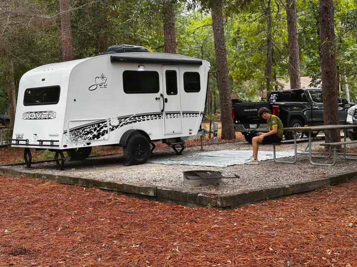 Enjoying an RV rental at a shaded campsite, featuring a white travel trailer, picnic table, & fire pit.. InTech RV Sol 2023
