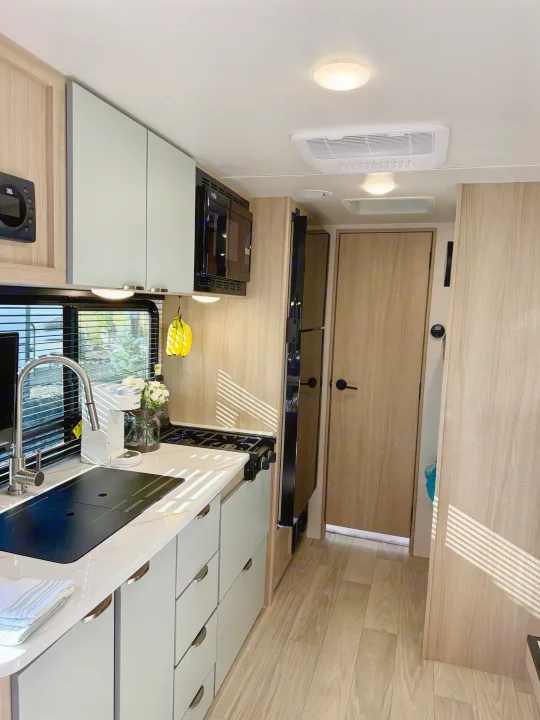 Bright, modern rv rental kitchen with light wood, blue-grey cabinets, sink, and cooktop.. Winnebago Micro Minnie 2025
