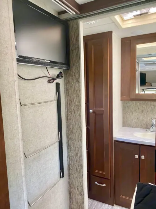 RV rental interior showing mounted TV, storage racks, and a compact bathroom with sink and dark wood cabinetry.. Forest River Sunseeker 2018
