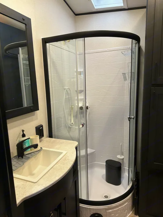 Modern RV rental bathroom: compact sink, black faucet, curved glass shower with white textured walls. Clean, functional.. Heartland Fuel 287 2022