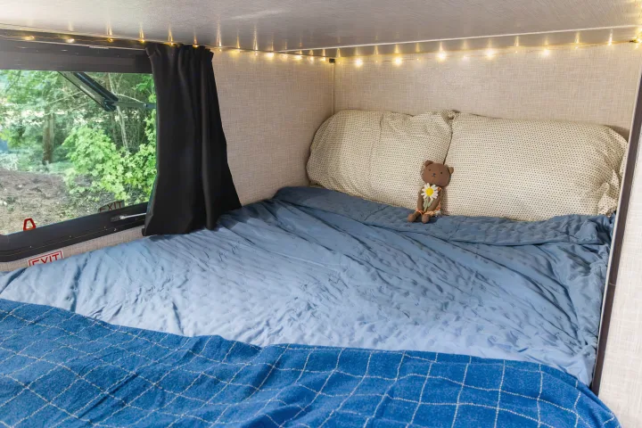 Experience the cozy bed in this RV rental, featuring string lights, a teddy bear, and a scenic forest window view.. Jayco Jay Flight SLX 2024