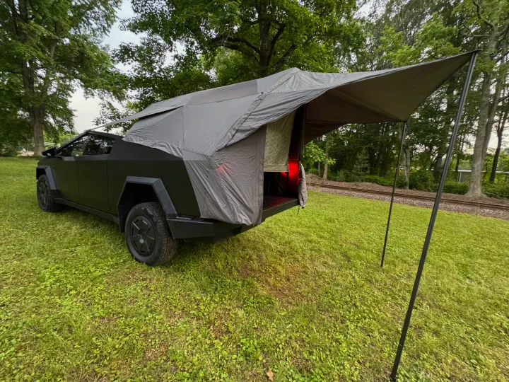 A Tesla Cybertruck, equipped with an expansive tent/awning, parked on grass near railroad tracks. Ideal for an electric RV rental experience.. Tesla Cybertruck 2025