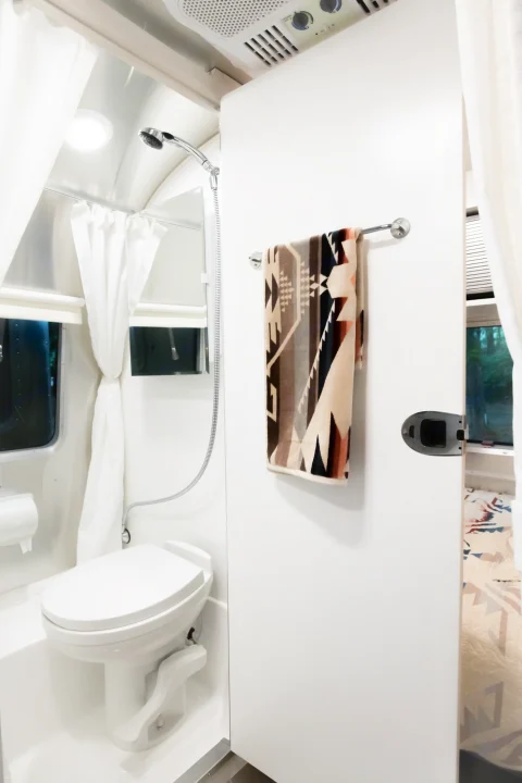 Inside an rv rental: a bright, compact white bathroom with a toilet and shower. A door to the living area holds a patterned towel.. Airstream Bambi 2022