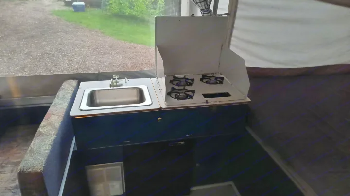 Pop-up RV rental interior: compact kitchen with sink, three-burner stove, and a view of the gravel outside.. Coleman Destiny Santa Fe 1996