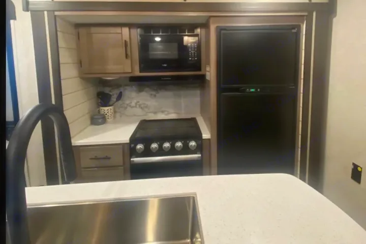 Modern RV rental kitchen with a stainless steel sink, black stove, microwave, and fridge, surrounded by light wood cabinets.. Crossroads RV Sunset Trail Super Lite 2023