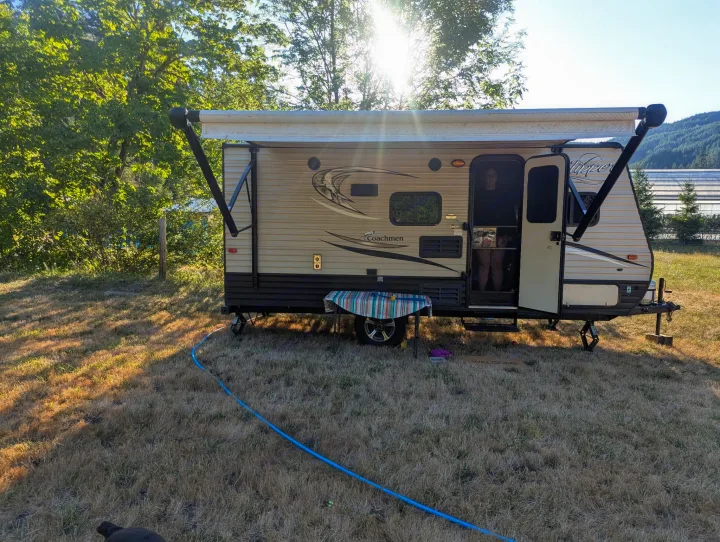 A tan RV rental travel trailer with an extended awning is parked on dry grass. A person is visible inside. Sunny trees and mountains in back.. Clipper 17BH 2018