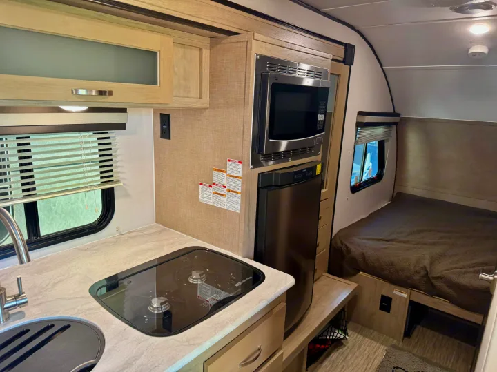 Inside this RV rental: a compact kitchenette with microwave, stovetop, sink, and fridge, plus a comfortable bed area.. Forest River RPod 189 2019