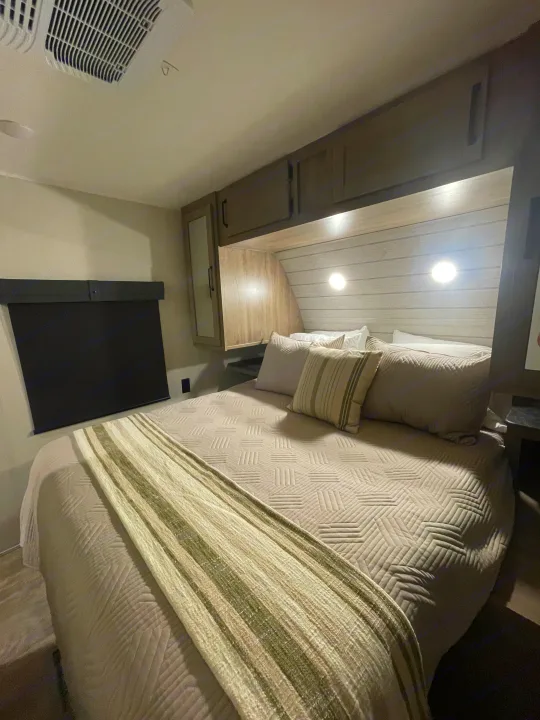 Cozy RV rental bedroom features a quilted bed, striped throw, overhead cabinetry, reading lights, and window blind.. Forest River Palomino Puma 2024
