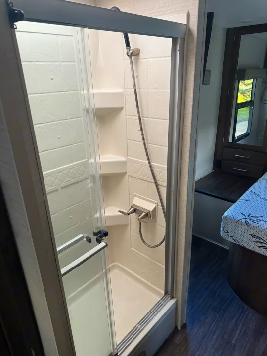 Clean, compact RV rental shower with glass doors and handheld shower head. Partial view of bedroom area.. Jayco Greyhawk 2018