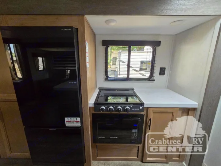 RV rental kitchen: modern black fridge, Contoure stove/oven, white counters, and a large window with blinds.. Forest River Cherokee M-274WK 2024