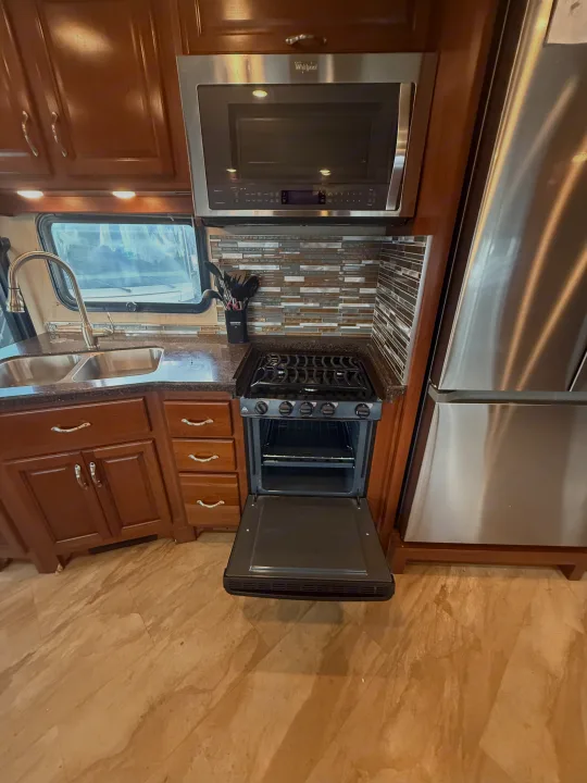 Modern rv rental kitchen featuring microwave, gas stove/oven, sink, and fridge, with wood cabinets & tile backsplash.. Fleetwood Bounder 2016