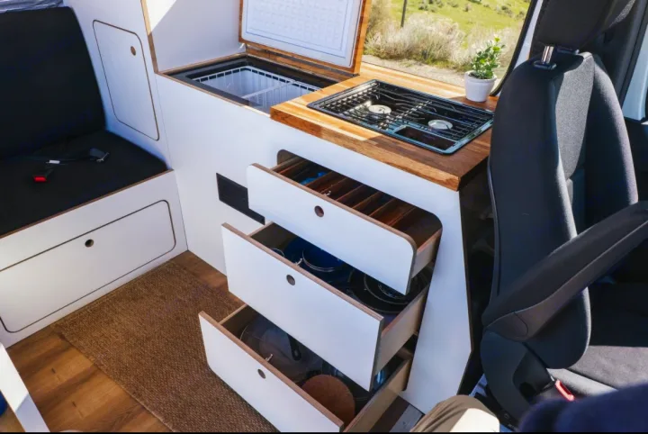 Bright RV rental interior with a compact kitchen: stovetop, top-loading fridge, and open drawers revealing kitchen items.. Mercedes-Benz Sprinter 2022