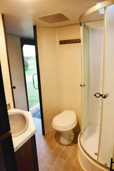Compact RV rental bathroom featuring a sink, toilet, and corner shower. An open door shows outdoor greenery.. Forest River Cherokee 2020