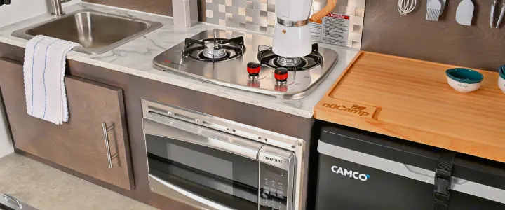 Fully equipped RV rental kitchen: stovetop, sink, Contoure microwave, and Camco cooler, plus nüCamp cutting board.. nuCamp T@G 2023