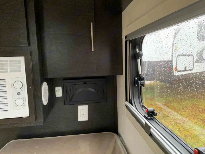 Cozy RV rental interior shows a window with rain outside, an AC unit, and dark cabinets. Ready for a rainy camp day.. nuCamp T@G 2023
