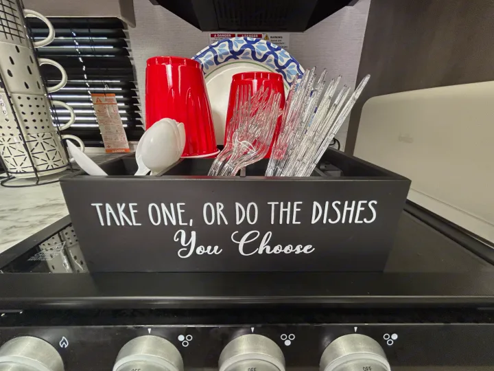 Humorous caddy in an rv rental kitchen: 'Take One, Or Do The Dishes,' with disposables & reusable mugs nearby.. Grand Design Transcend Xplor 2024