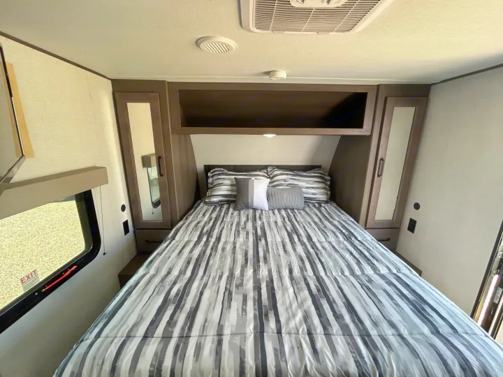 Explore this cozy RV rental bedroom interior, featuring a striped bed, overhead cabinets, and built-in storage on both sides.. Grand Design Transcend Xplor 2024