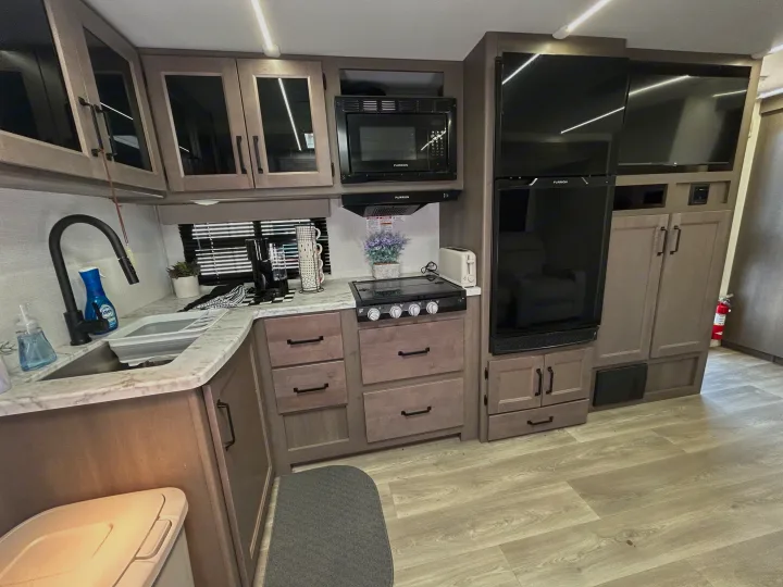 Modern RV rental kitchen featuring a microwave, stove, sink, refrigerator, and TV within light wood cabinets and stone-look counters.. Grand Design Transcend Xplor 2024