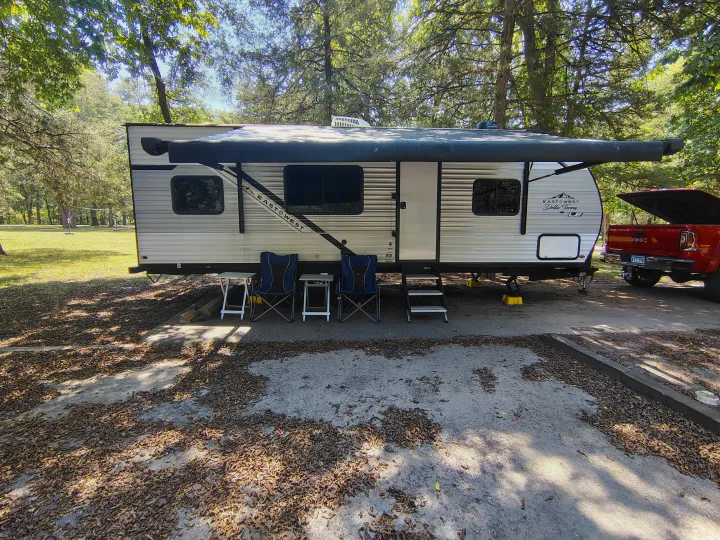 RV rental: a travel trailer with an awning extended, chairs, and a red pickup truck at a shaded, leafy campsite.. East to West Della Terra LE 2024