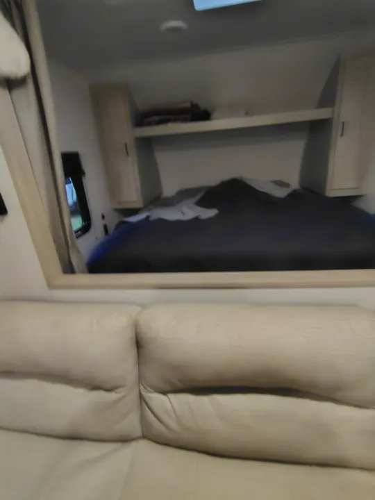 View of an RV rental bedroom showing a made bed, overhead storage, a small window, and a light-colored sofa below.. East to West Della Terra LE 2024